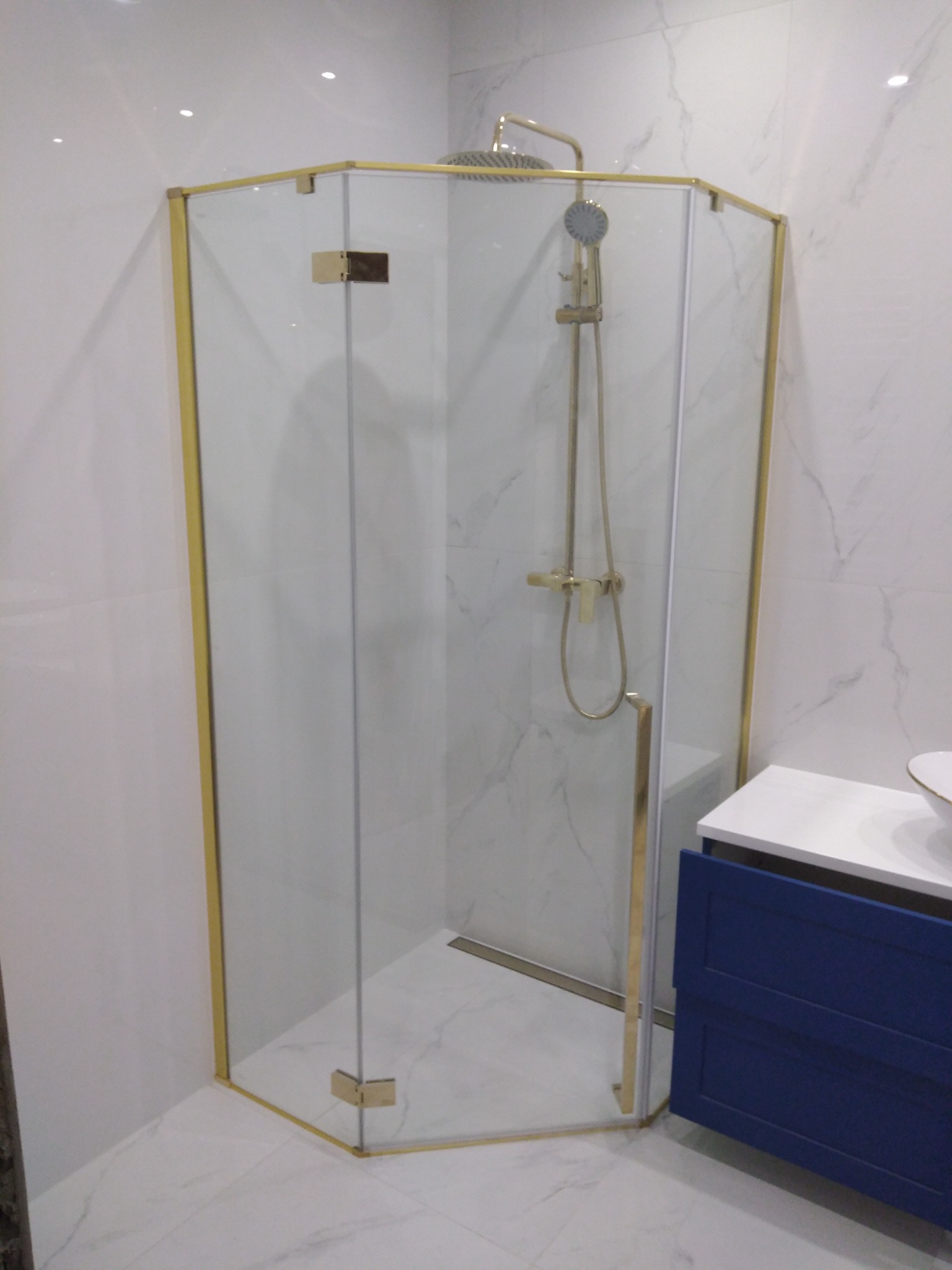 Nowoczesna kabina prysznicowa z ośmiokątną szybą i złotymi profilami, dopasowana kolorystycznie do armatury, na tle białych płytek imitujących marmur i granatowej szafki umywalkowej.