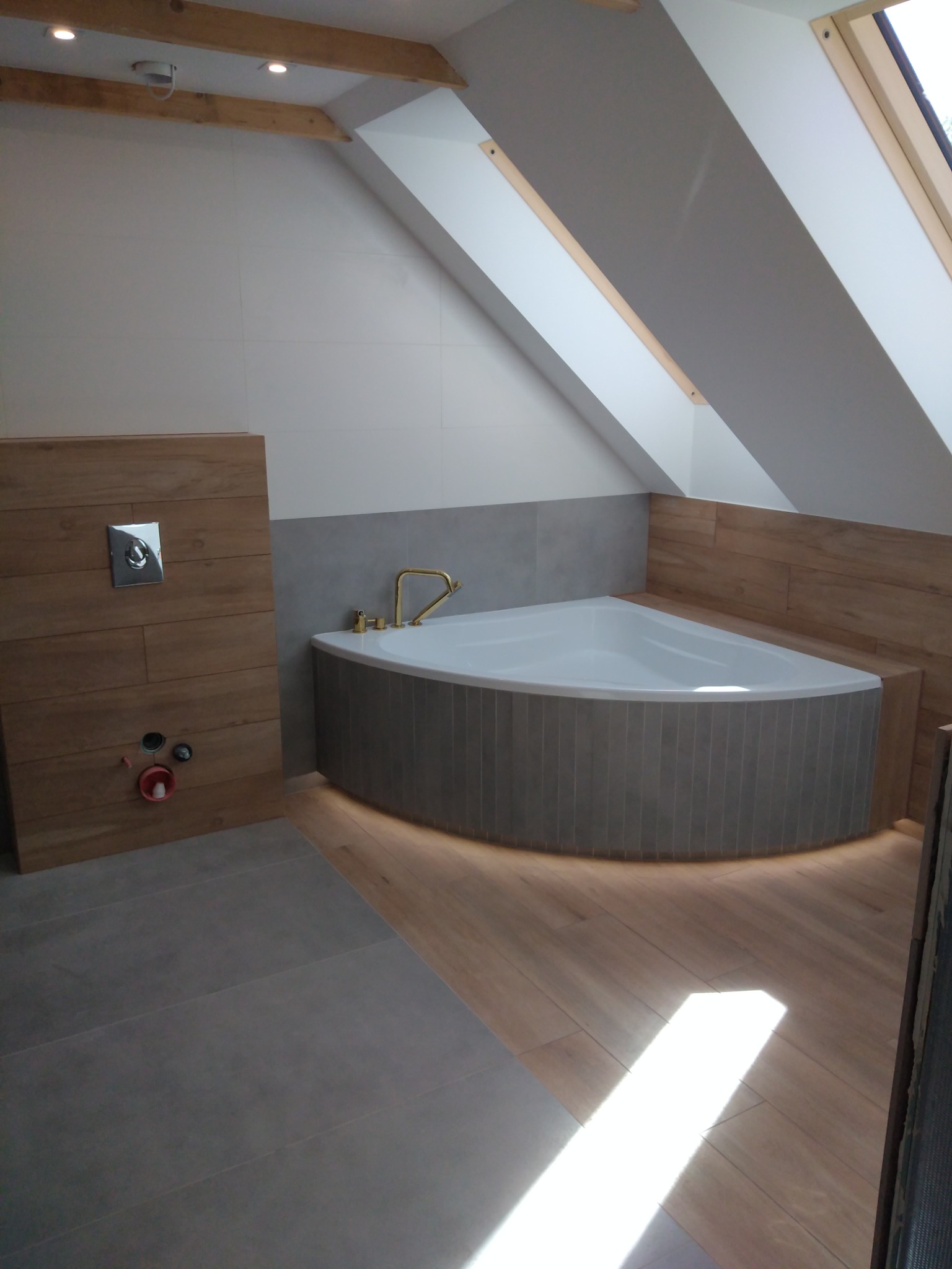 Nowoczesna łazienka na poddaszu ze skośnym oknem dachowym, narożną wanną z podświetleniem LED, drewnopodobnymi płytkami na ścianie i podłodze oraz szarą posadzką z płytek.