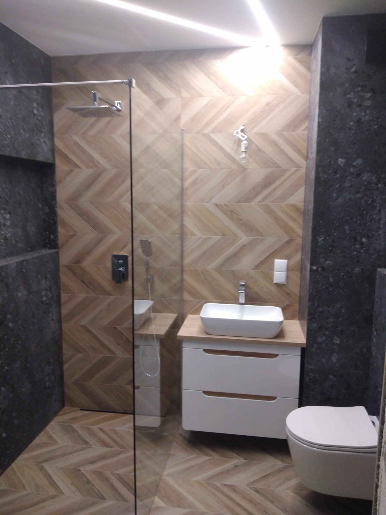 Nowoczesna łazienka z kabiną prysznicową typu walk-in, umywalką nablatową na białej szafce z drewnianym blatem i podwieszaną toaletą, wykończona płytkami w jodełkę i ciemnym kamieniem.