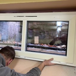 BUD-EXPERT AGATA KULCZYCKA - Białe dwuskrzydłowe okno podczas montażu w surowym otworze okiennym, widoczne fragmenty muru i izolacji, instalator w szarej bluzie przytrzymuje ramę.