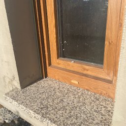 BUD-EXPERT AGATA KULCZYCKA - Zamontowany parapet z konglomeratu o szaro-beżowej fakturze pod oknem PCV o drewnopodobnej ramie, widoczne ślady prac montażowych.