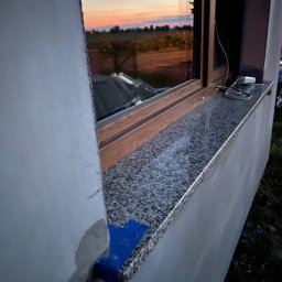 BUD-EXPERT AGATA KULCZYCKA - Montaż okna z parapetem z konglomeratu, widoczny fragment ściany w trakcie wykończenia, zarys zachodzącego słońca odbity w szybie.