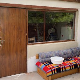 BUD-EXPERT AGATA KULCZYCKA - Brązowe drzwi zewnętrzne z folią ochronną na klamce, okno z drewnianą ramą i fragment sofy na tle białej ściany budynku.