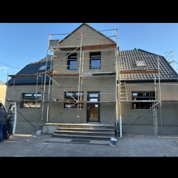 BUD-EXPERT AGATA KULCZYCKA - Dom w trakcie ocieplania, z widocznymi rusztowaniami, szarą elewacją, czarnymi ramami okiennymi i drzwiami oraz częściowo widocznym samochodem dostawczym.