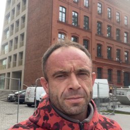 BUD-EXPERT AGATA KULCZYCKA - Mężczyzna w czerwonej bluzie moro na tle odnowionej kamienicy z nowymi oknami, widoczne samochody dostawcze i barierki budowlane.