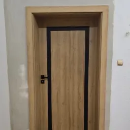 Zamontowane drzwi wewnętrzne z drewnianą ramą i czarnym obramowaniem na tle ściany w trakcie wykańczania. Widoczny fragment podłogi.