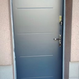 BUD-EXPERT AGATA KULCZYCKA - Grafitowe drzwi zewnętrzne z trzema poziomymi frezami, srebrną klamką i kluczami w zamku, osadzone w ościeżnicy z jasną fugą, na tle beżowej elewacji.