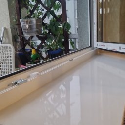 BUD-EXPERT AGATA KULCZYCKA - Nowy, błyszczący parapet wewnętrzny z widocznym fragmentem ramy okiennej, balkonem z roślinami w tle i fragmentem grzejnika pod spodem.