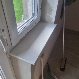 BUD-EXPERT AGATA KULCZYCKA - Montaż białego parapetu z tworzywa, widoczne niedociągnięcia w wykończeniu ściany, obok stoi zmiotka i szufelka.