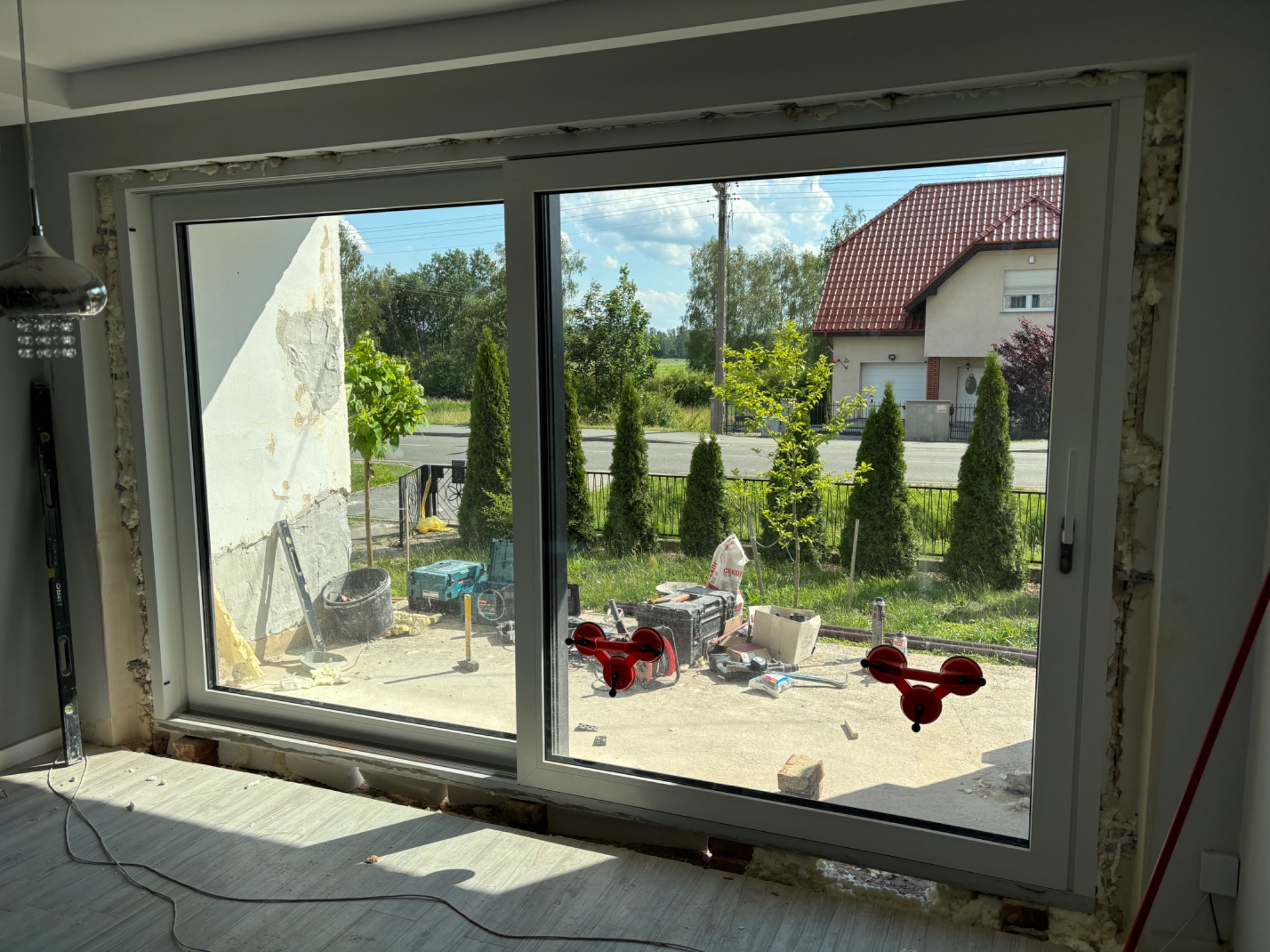 Montaż dużego okna przesuwnego w Poniatowicach, widoczne narzędzia, piana montażowa i przyssawki do szyb, widok na dom z czerwonym dachem.