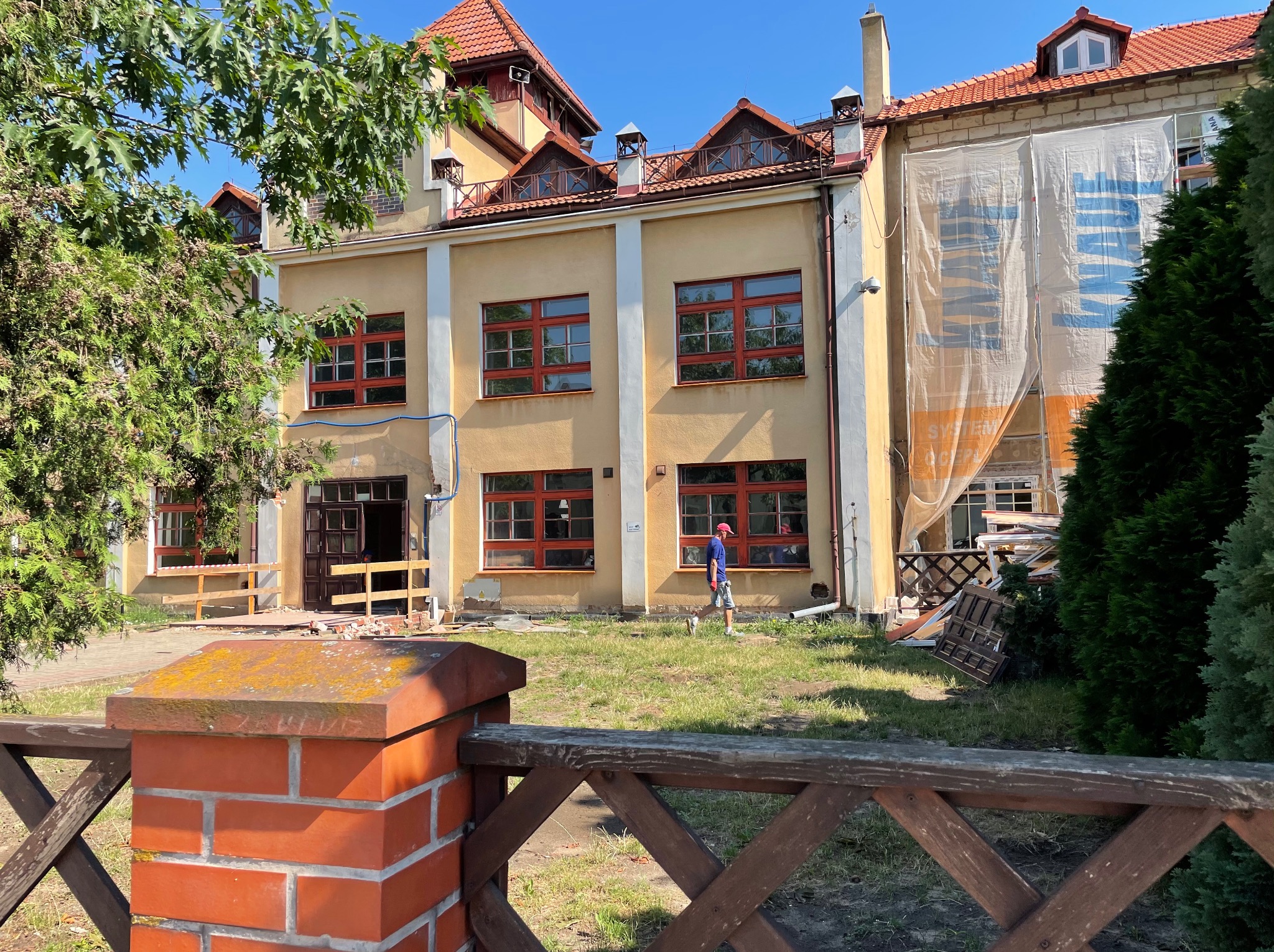 Budynek w trakcie remontu: widoczne rusztowania, okna z czerwonymi ramami, częściowo zasłonięta elewacja, pracownik w oddali i elementy demontażu na trawniku.