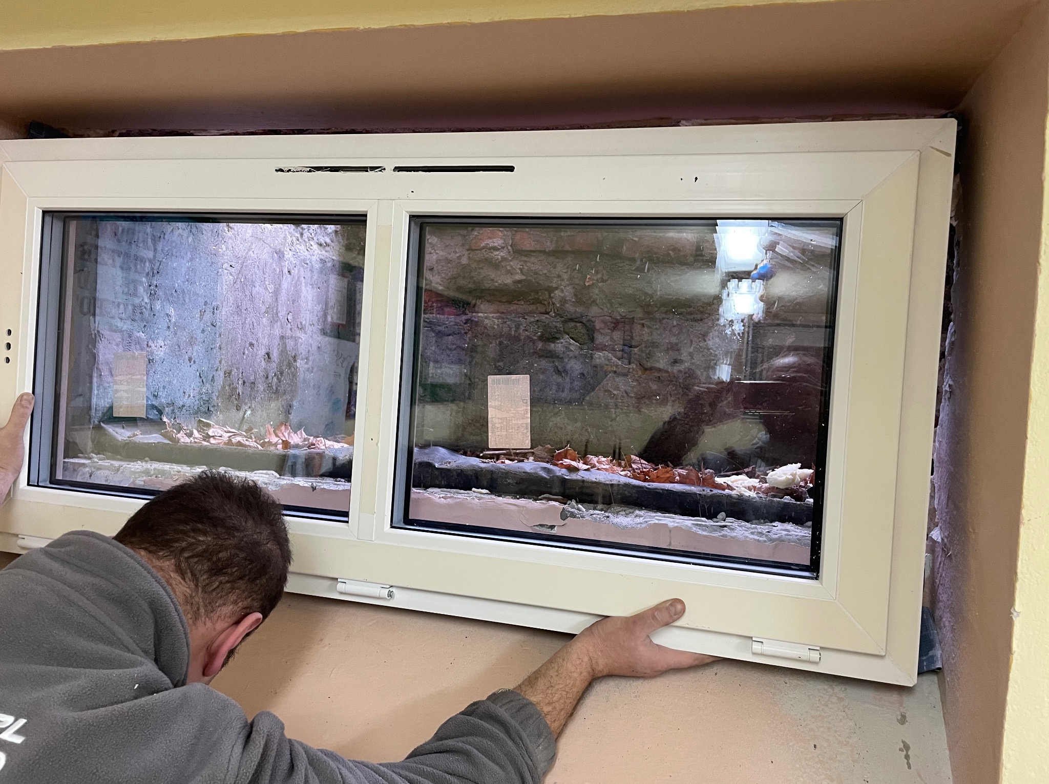 Białe dwuskrzydłowe okno podczas montażu w surowym otworze okiennym, widoczne fragmenty muru i izolacji, instalator w szarej bluzie przytrzymuje ramę.
