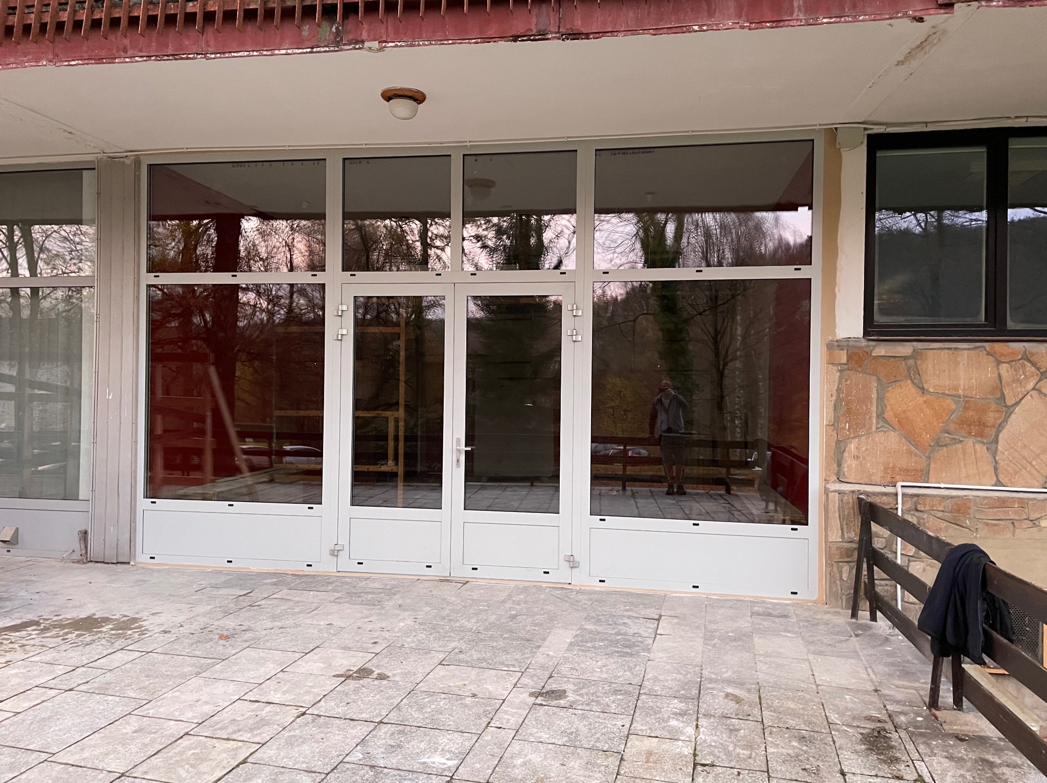 Białe drzwi balkonowe z aluminiową ramą, widoczne odbicie osoby i otoczenia w szybach, szara kamienna posadzka tarasu.