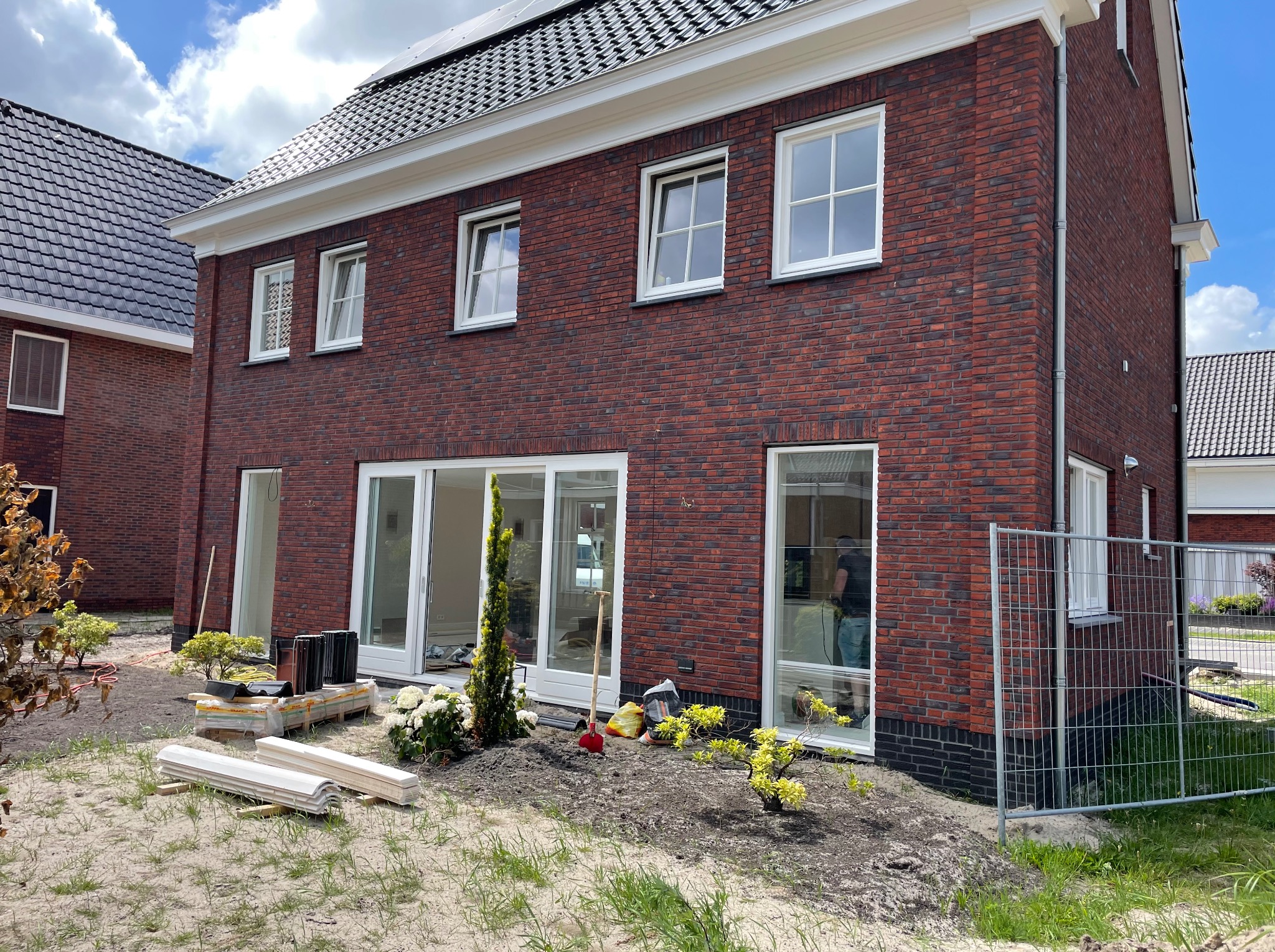 Nowo wybudowany dom z czerwonej cegły z białymi ramami okiennymi i przesuwnymi drzwiami tarasowymi, widoczne materiały budowlane sugerują prace wykończeniowe wokół budynku.