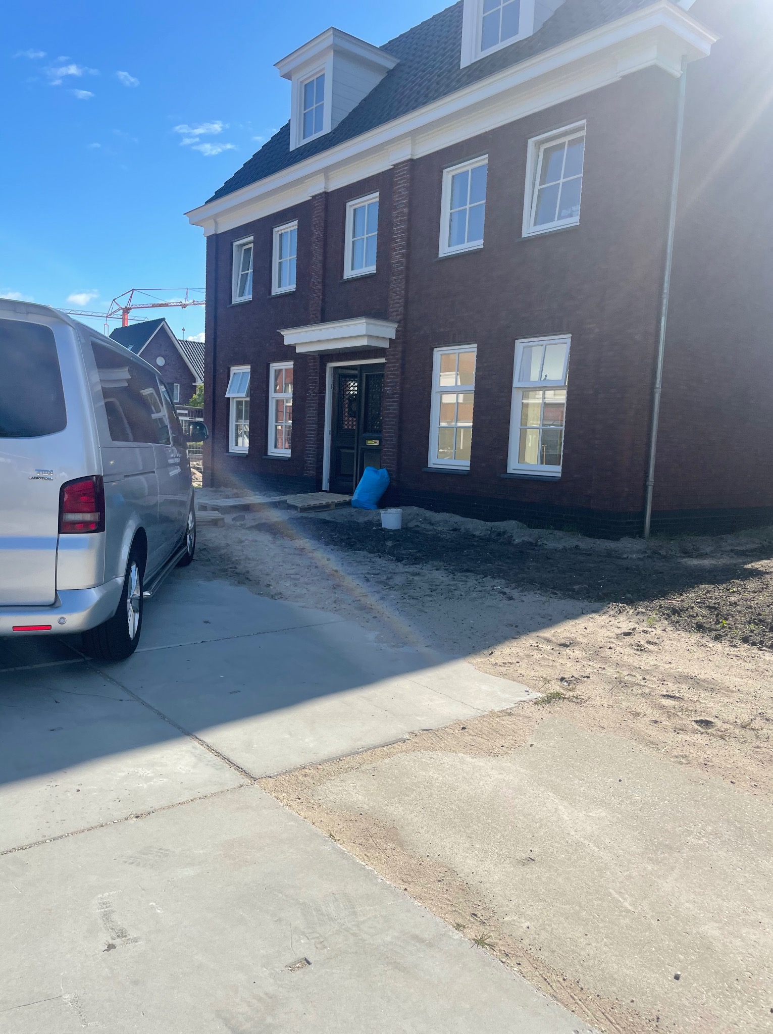 Nowo wybudowany dom z cegły z białymi oknami PCV i aluminiowym minivanem zaparkowanym na betonowym podjeździe, widoczny żuraw budowlany w tle.