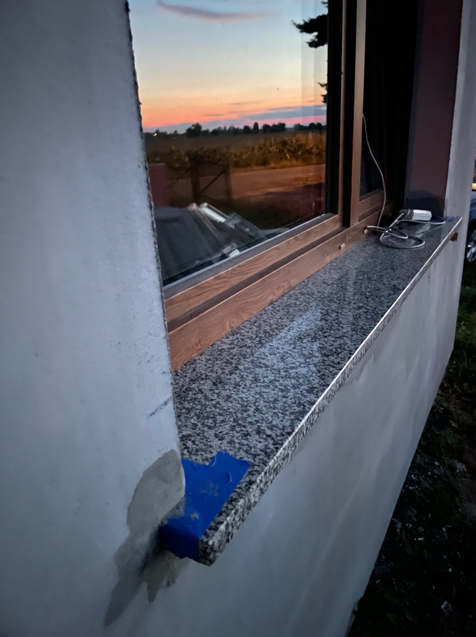 Montaż okna z parapetem z konglomeratu, widoczny fragment ściany w trakcie wykończenia, zarys zachodzącego słońca odbity w szybie.