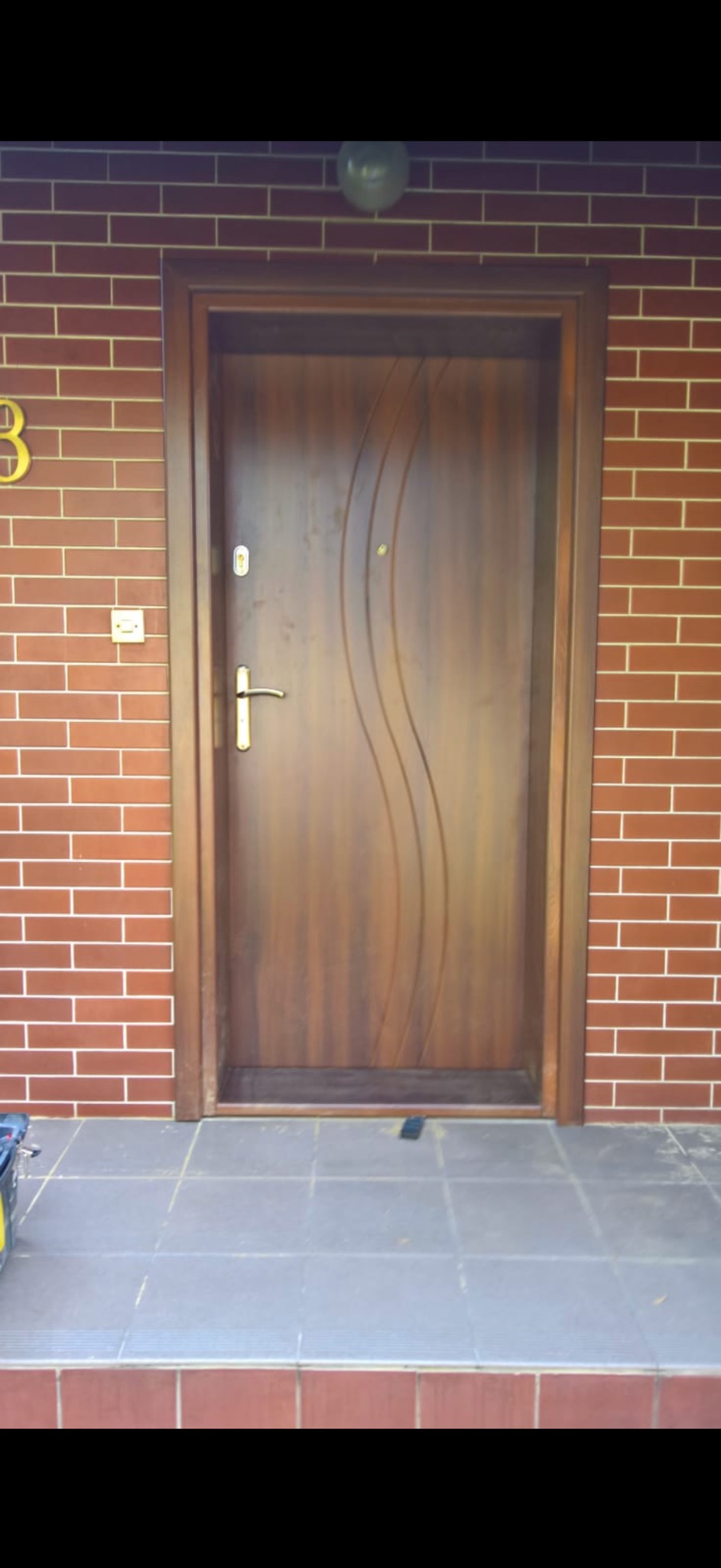 Brązowe drzwi wejściowe z dekoracyjnym, falistym frezem, zamontowane w ścianie z cegły klinkierowej i złotym numerem domu 3, widoczne z poziomu podłogi wyłożonej szarymi płytkami.