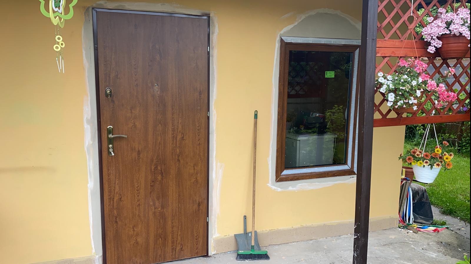 Brązowe drzwi wejściowe z ościeżnicą, świeżo zamontowane w żółtej ścianie budynku, widoczna szpachelka i szczotka do zamiatania, okno z brązową ramą i kwiaty w donicach na zewnątrz.