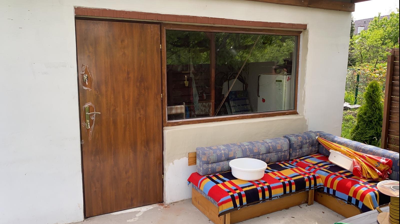 Brązowe drzwi zewnętrzne z folią ochronną na klamce, okno z drewnianą ramą i fragment sofy na tle białej ściany budynku.