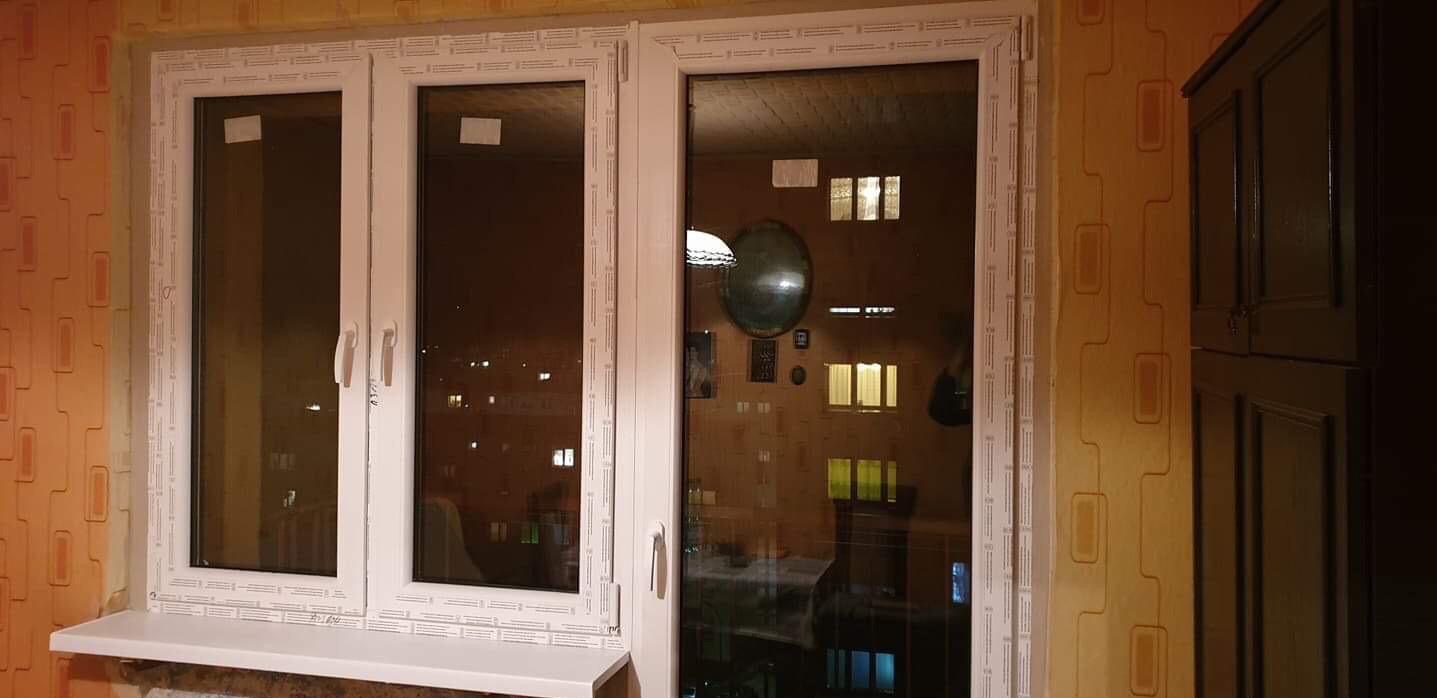 Trzyczęściowe okno PCV z białymi ramami i parapetem, widok na oświetlone nocne miasto przez szybę.