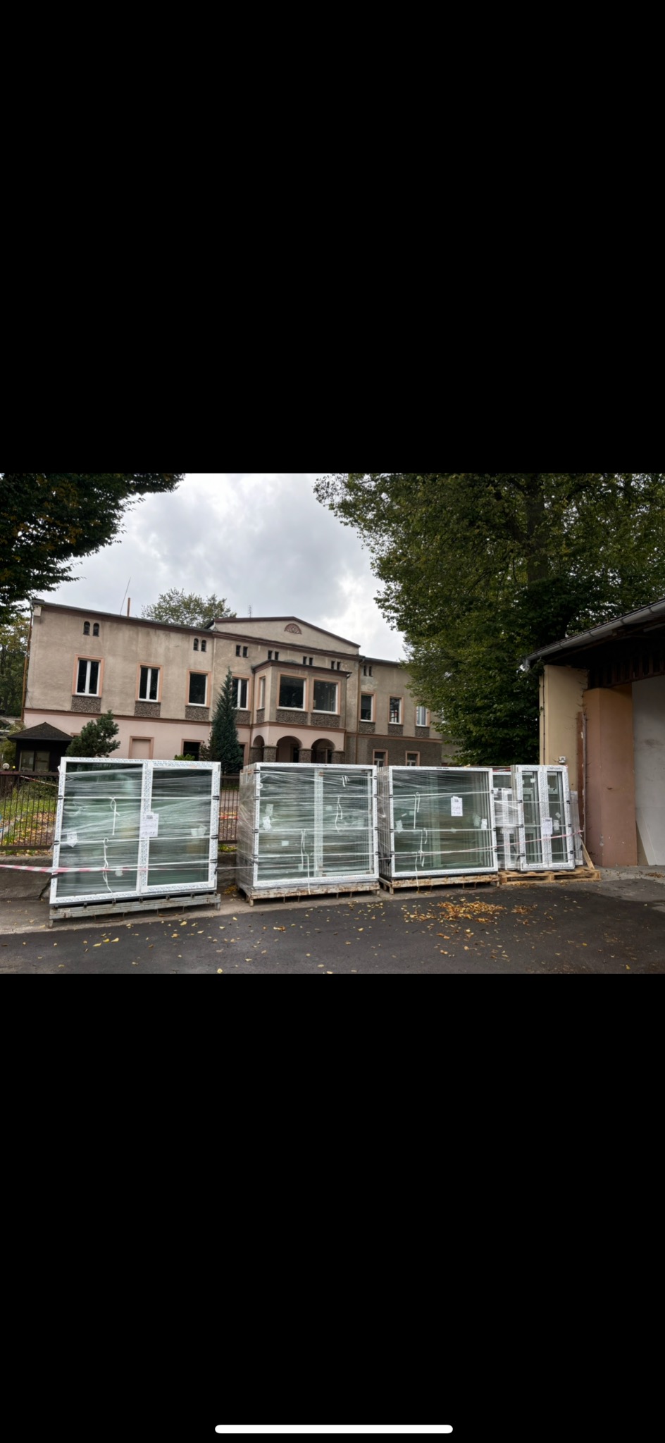 Pakiety okien z białymi ramami, zabezpieczone folią, składowane na paletach przed budynkiem z beżową elewacją i drzewami w tle.