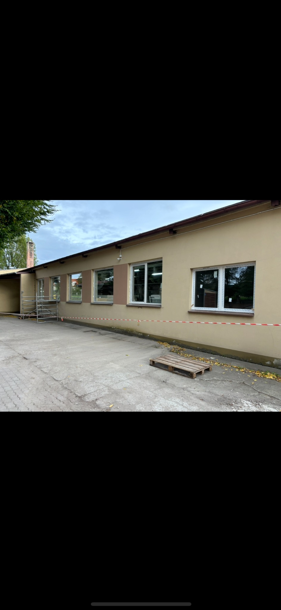 Budynek z beżową elewacją po wymianie okien, widoczne nowe białe ramy okienne, zabezpieczone taśmą ostrzegawczą i rusztowanie przy ścianie bocznej.