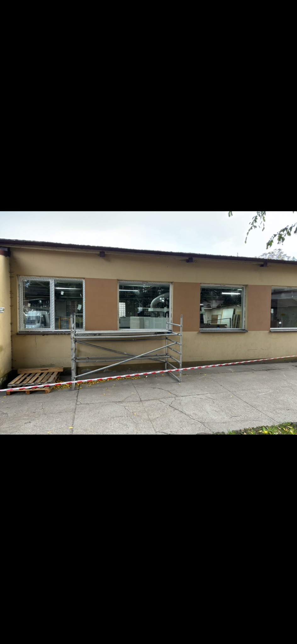 Budynek z zamontowanymi oknami PCV, widoczne rusztowanie i taśma ostrzegawcza, paleta drewniana obok