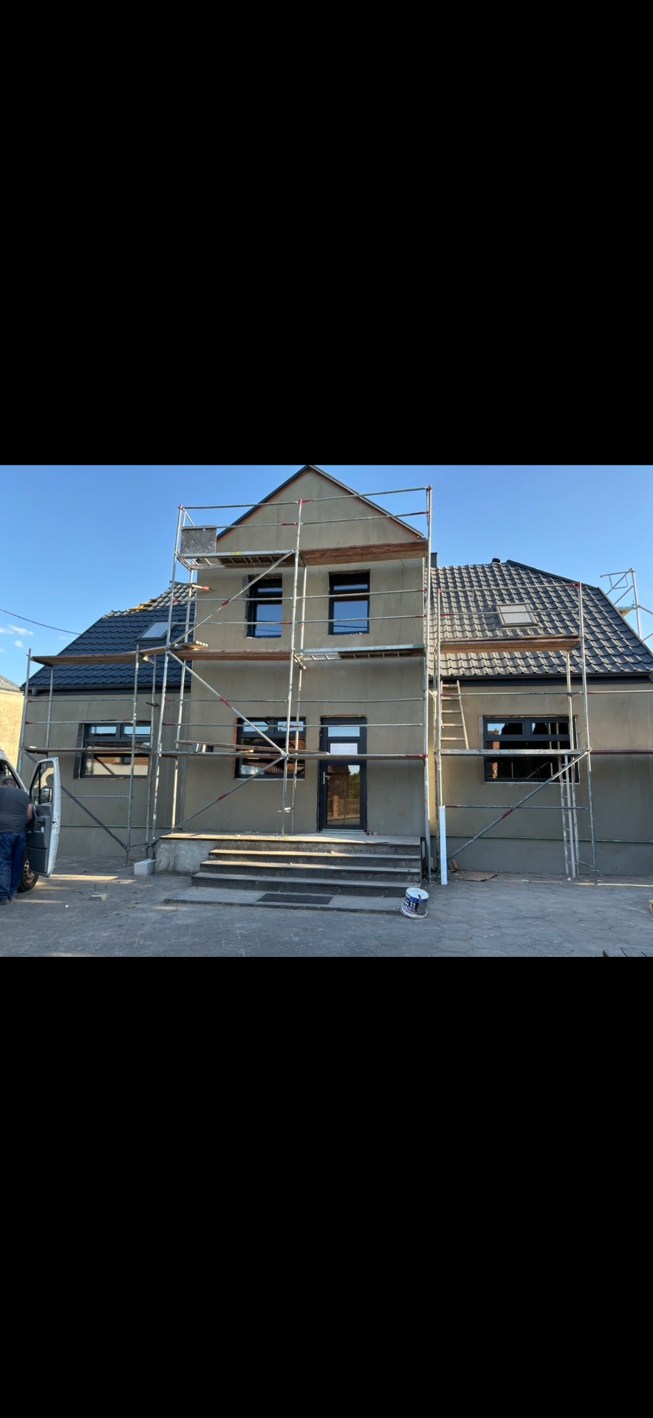 Dom w trakcie ocieplania, z widocznymi rusztowaniami, szarą elewacją, czarnymi ramami okiennymi i drzwiami oraz częściowo widocznym samochodem dostawczym.