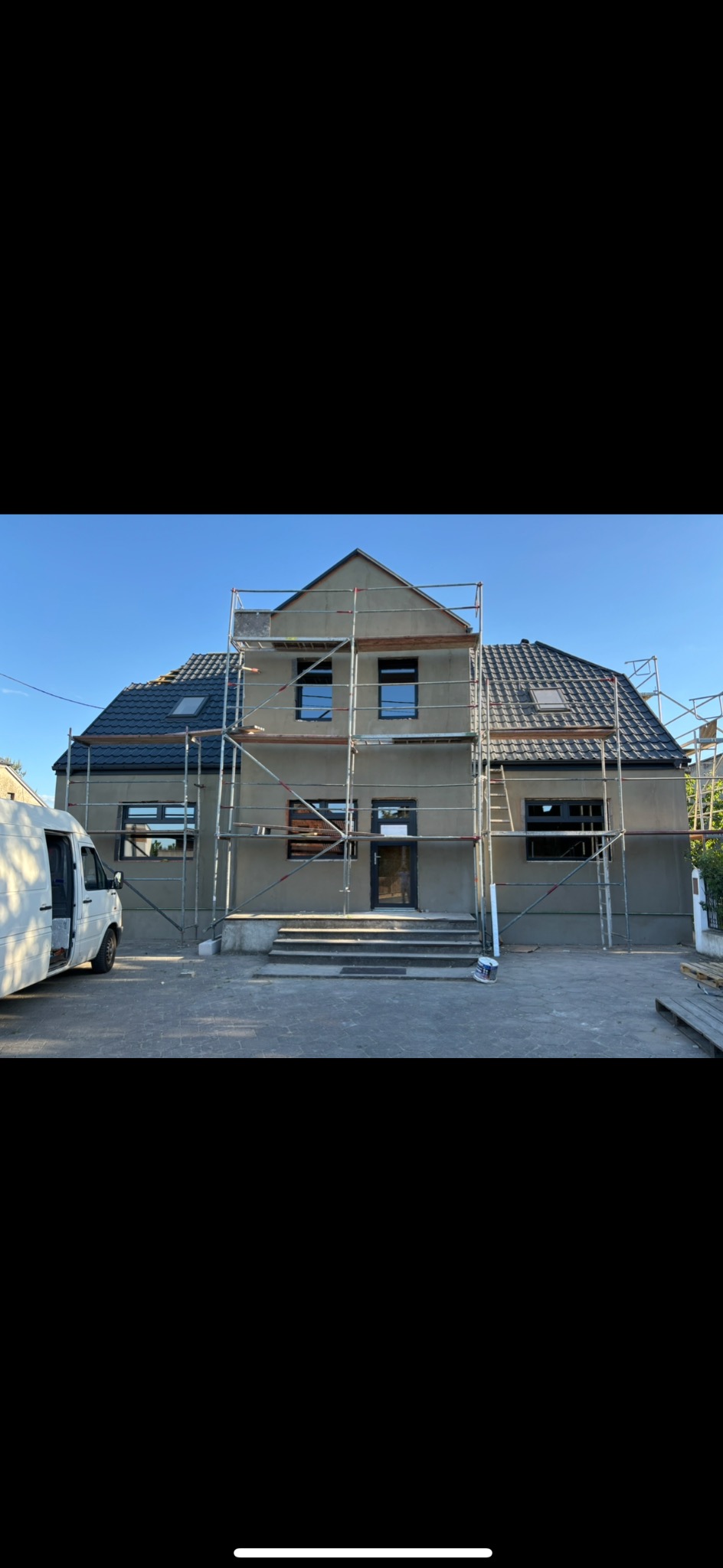 Dom jednorodzinny w trakcie prac elewacyjnych, widoczne rusztowania, zamontowane okna, świeżo otynkowana fasada w szarym odcieniu oraz częściowo wykończone schody wejściowe.