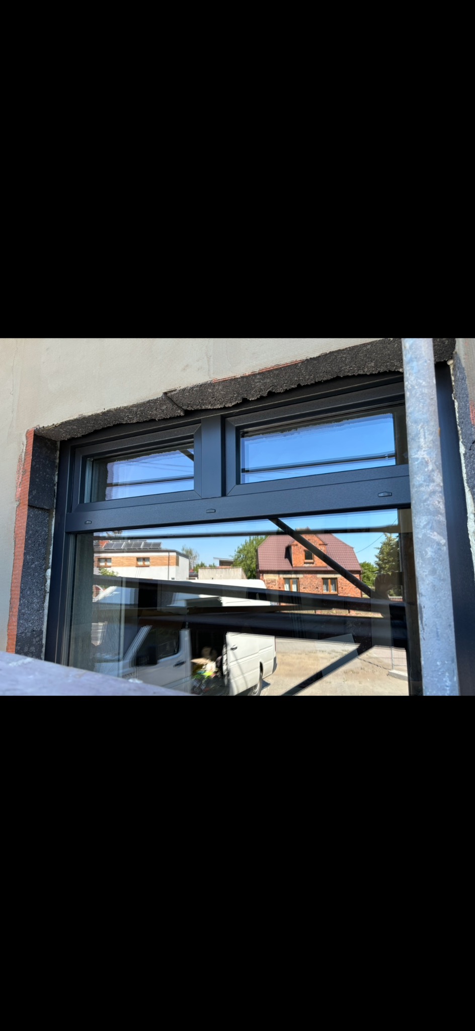 Montaż czarnego okna z dwiema szybami, widoczne przygotowane ocieplenie ściany i odbicie białego samochodu dostawczego w szybie.