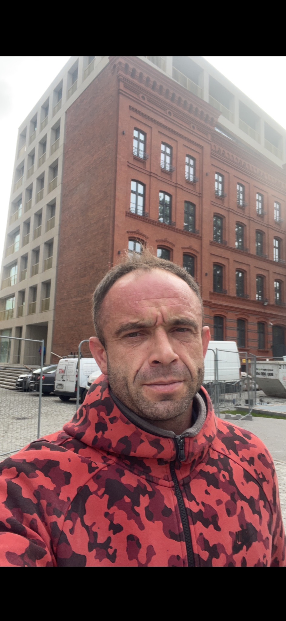 Mężczyzna w czerwonej bluzie moro na tle odnowionej kamienicy z nowymi oknami, widoczne samochody dostawcze i barierki budowlane.