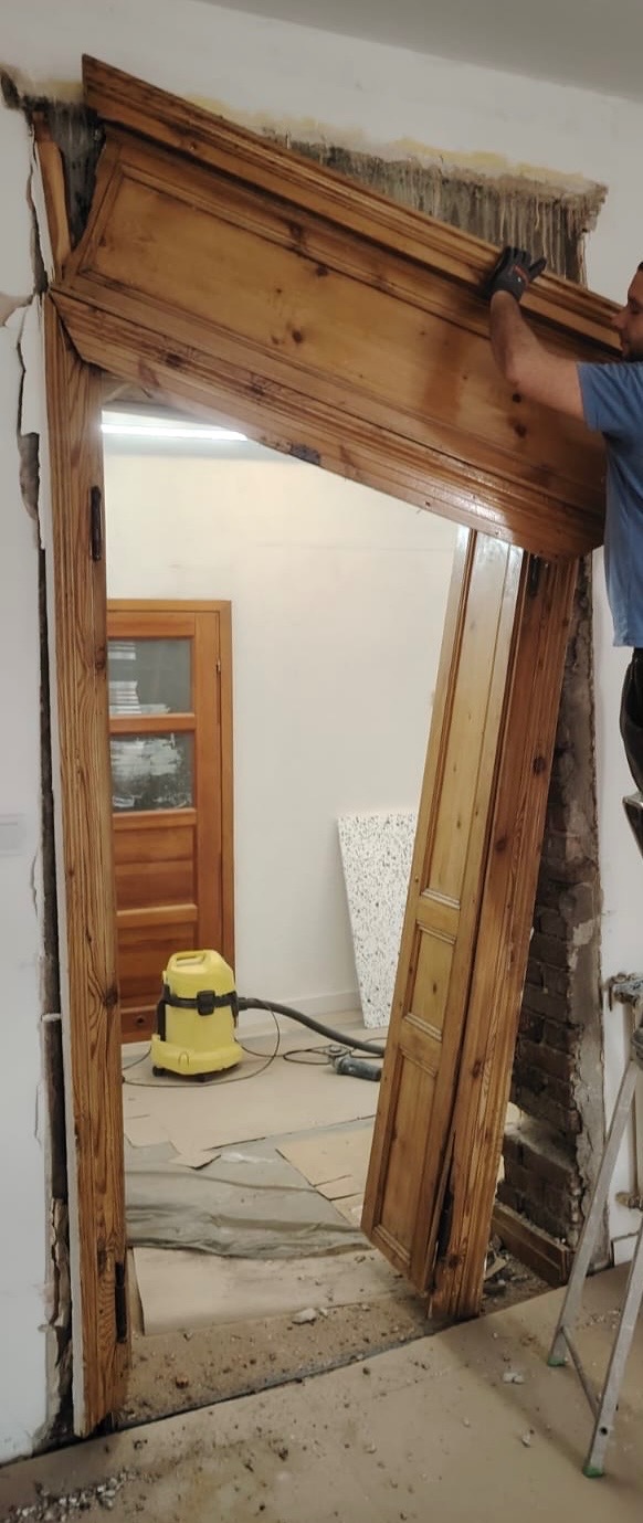 Demontaż drewnianej ościeżnicy drzwiowej podczas remontu. Widoczne uszkodzenia ściany i przygotowane podłoże. W tle widoczny odkurzacz przemysłowy.
