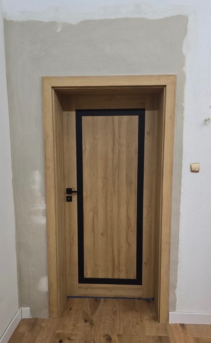 Zamontowane drzwi wewnętrzne z drewnianą ramą i czarnym obramowaniem na tle ściany w trakcie wykańczania. Widoczny fragment podłogi.