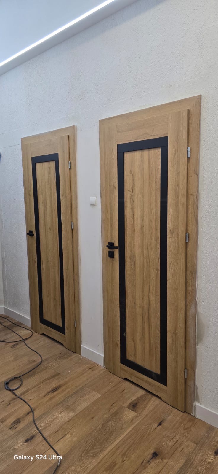 Dwa identyczne, drewniane drzwi wewnętrzne z czarnymi ramkami, zamontowane w białej ścianie. Widoczne kable na podłodze wskazują na świeży montaż.