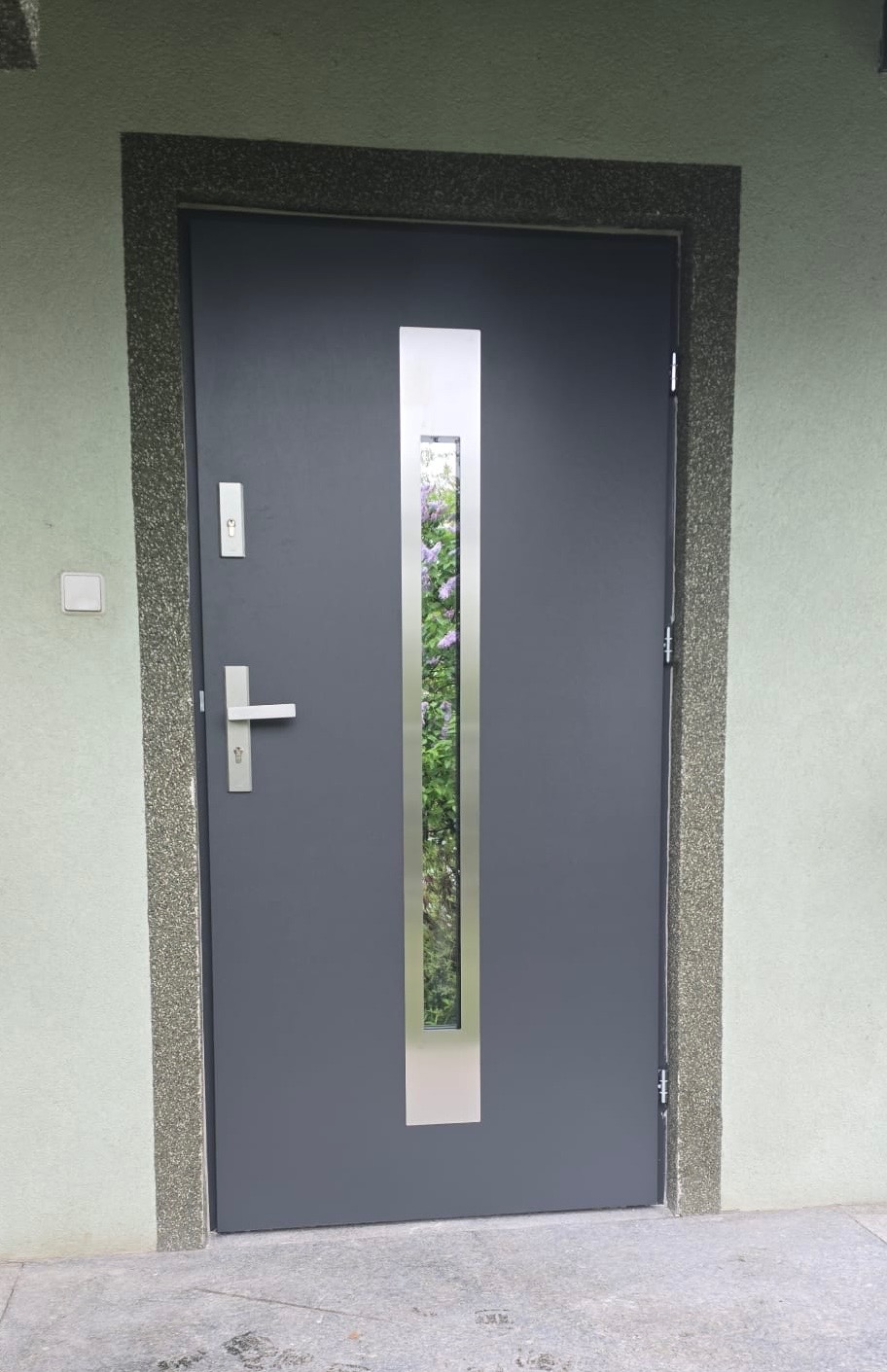 Ciemnoszare drzwi wejściowe z podłużnym przeszkleniem, przez które widać fioletowe kwiaty. Drzwi osadzone w ościeżnicy z szorstkiego, kamiennego materiału.