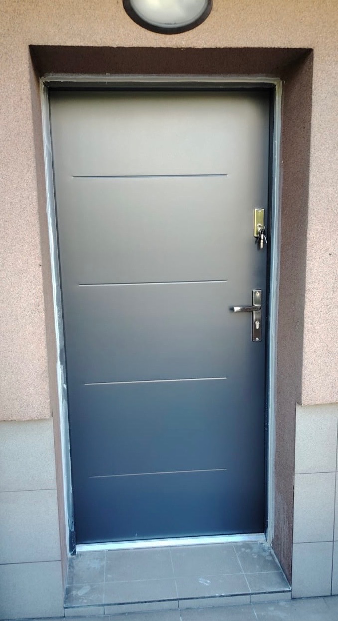 Grafitowe drzwi zewnętrzne z trzema poziomymi frezami, srebrną klamką i kluczami w zamku, osadzone w ościeżnicy z jasną fugą, na tle beżowej elewacji.
