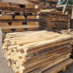 Stosy ułożonych, świeżo przetartych desek i półokrąglaków z korą, czekających na obróbkę lub sprzedaż, widoczne na paletach.