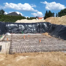 Przygotowanie zbrojenia pod płytę fundamentową z widocznymi pionowymi rurami instalacyjnymi, zabezpieczone czarną folią, w tle usypana ziemia i zabudowa jednorodzinna.