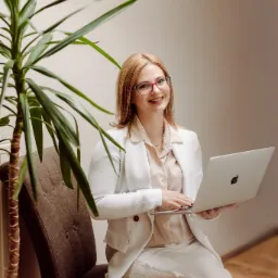 Uśmiechnięta kobieta w białym garniturze i okularach siedzi na krześle z laptopem Apple, obok dużej rośliny doniczkowej.