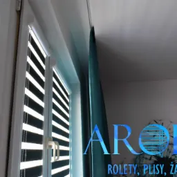 Fragment okna z zamontowaną przesłoną okienną typu dzień i noc z widocznymi poziomymi pasami w kolorze białym i czarnym, obok zasłona w kolorze morskim i logo firmy na ścianie.