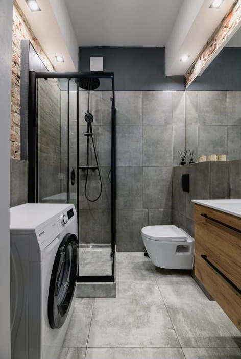 Nowoczesna łazienka z kabiną prysznicową typu walk-in z czarnymi ramami, pralką, toaletą wiszącą i szafką z drewnianym frontem, wykończona szarymi płytkami imitującymi beton.