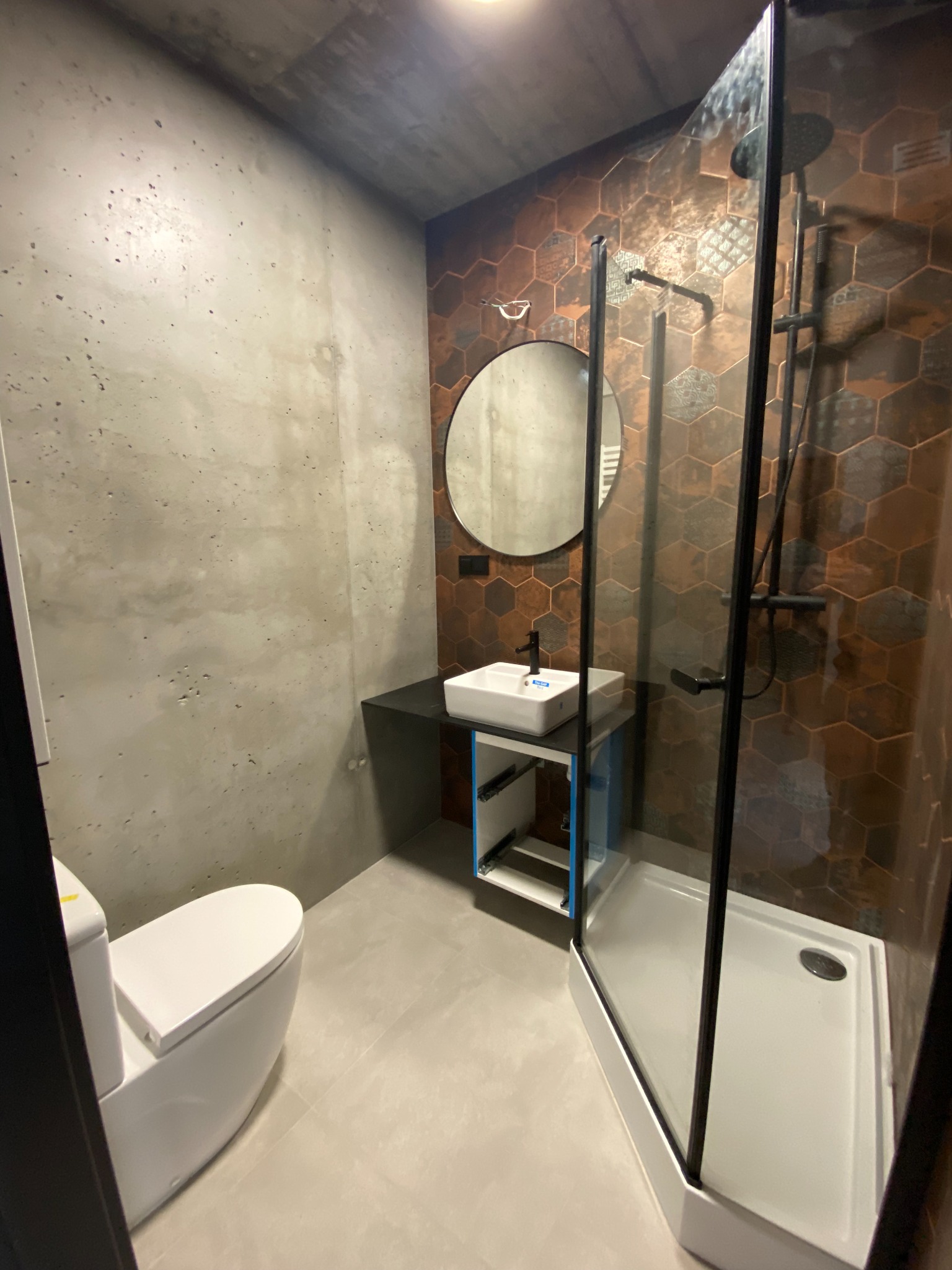 Nowoczesna łazienka z betonową ścianą, heksagonalnymi płytkami w odcieniach brązu, kabiną prysznicową z czarną armaturą, okrągłym lustrem i białą umywalką na czarnym blacie.