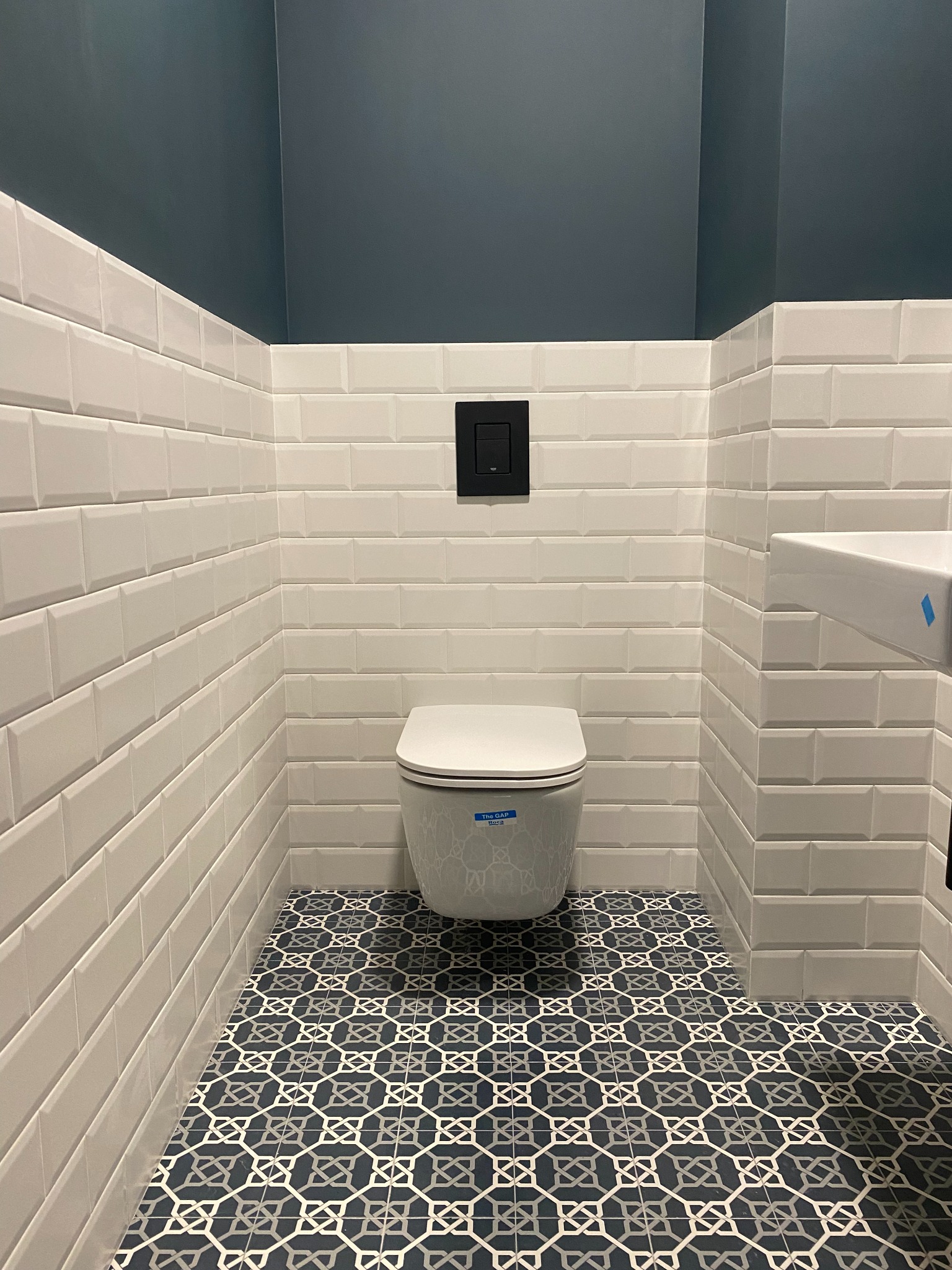 Nowoczesna łazienka z białymi, prostokątnymi płytkami ściennymi imitującymi cegłę, dekoracyjną, ciemną podłogą w geometryczne wzory oraz podwieszaną toaletą z czarnym przyciskiem spłukującym.