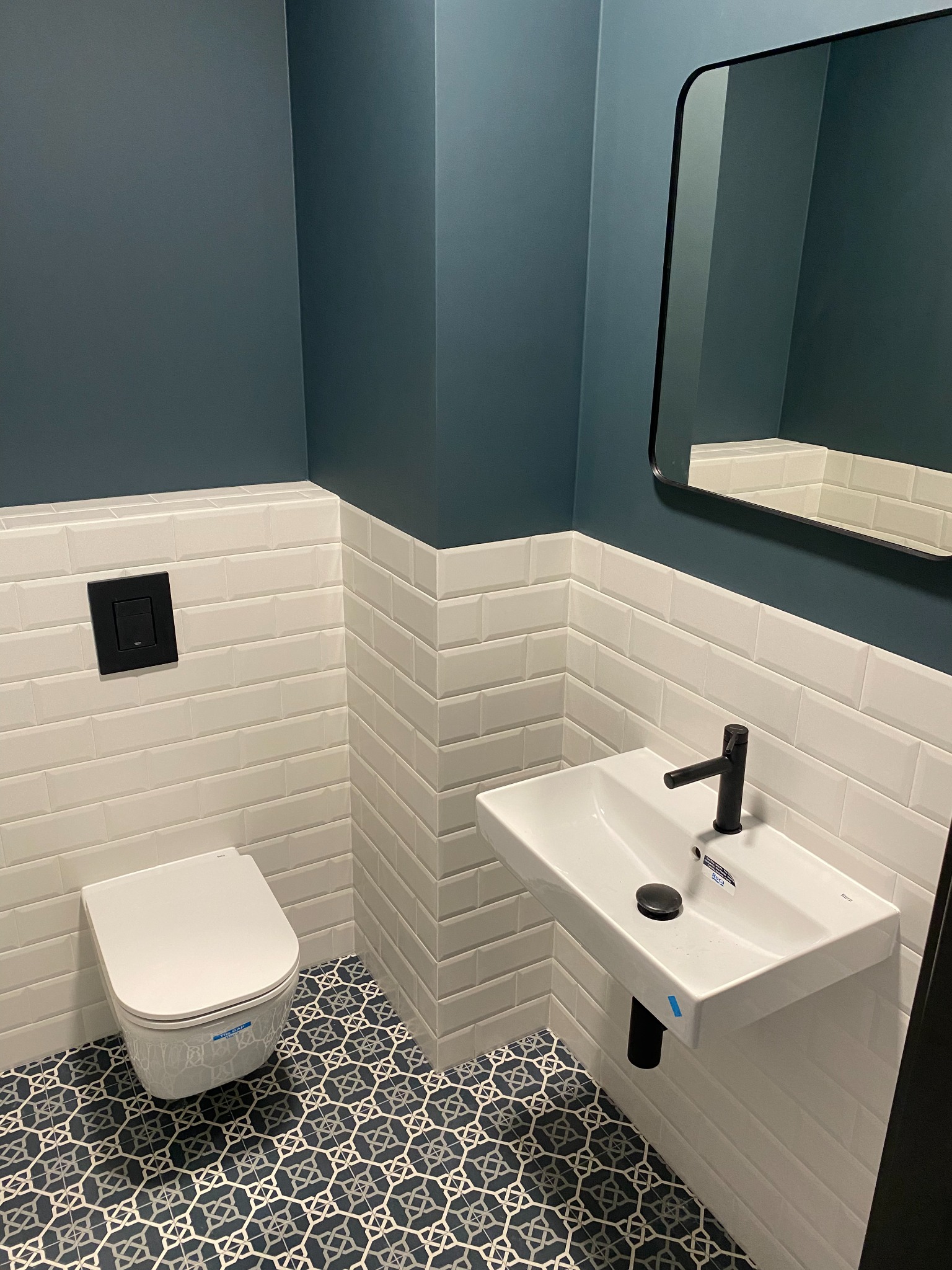 Nowoczesna łazienka z wiszącą toaletą, umywalką z czarną armaturą i geometryczną podłogą w odcieniach granatu i bieli, wykończona białymi płytkami typu metro do połowy wysokości ścian.