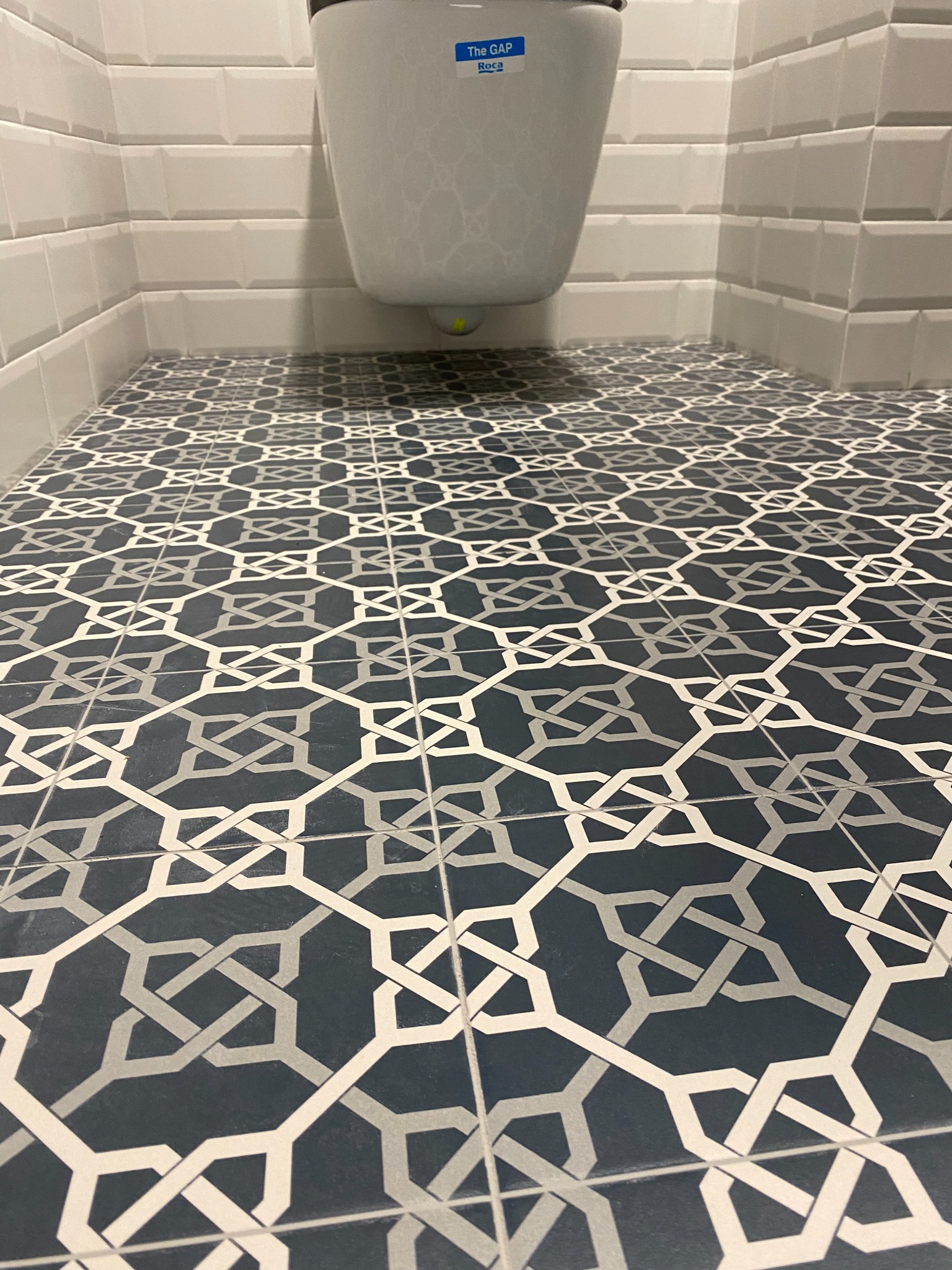 Wzorzysta, ciemna podłoga z płytek ceramicznych w łazience, widoczna pod wiszącą toaletą Roca i przy białych, prostokątnych płytkach ściennych ułożonych w cegiełkę.