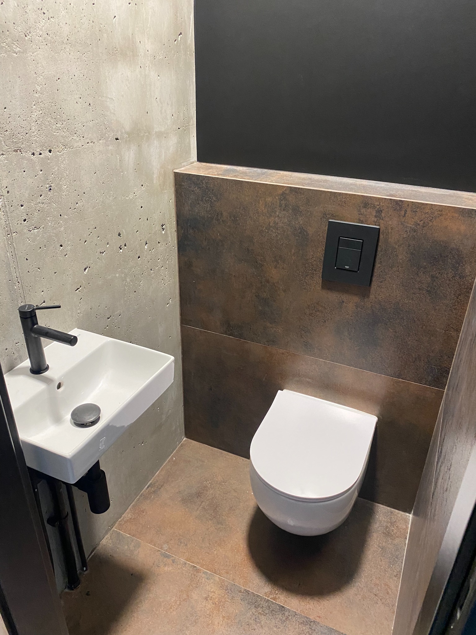 Nowoczesna, minimalistyczna toaleta: wisząca miska WC, umywalka z czarną armaturą, ściany wykończone betonem architektonicznym i płytkami imitującymi rdzę, czarny panel spłuczki.