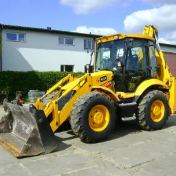 Żółta koparko-ładowarka JCB 3CX z brudną łyżką na kostce betonowej, w tle budynek z żywopłotem i przyczepa transportowa.