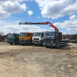 Flota ciężarówek MAN i Mercedes z dźwigiem HMF 1250, stojąca na placu budowy z gruzem i pociętymi kłodami drewna w tle, pod błękitnym niebem z obłokami.