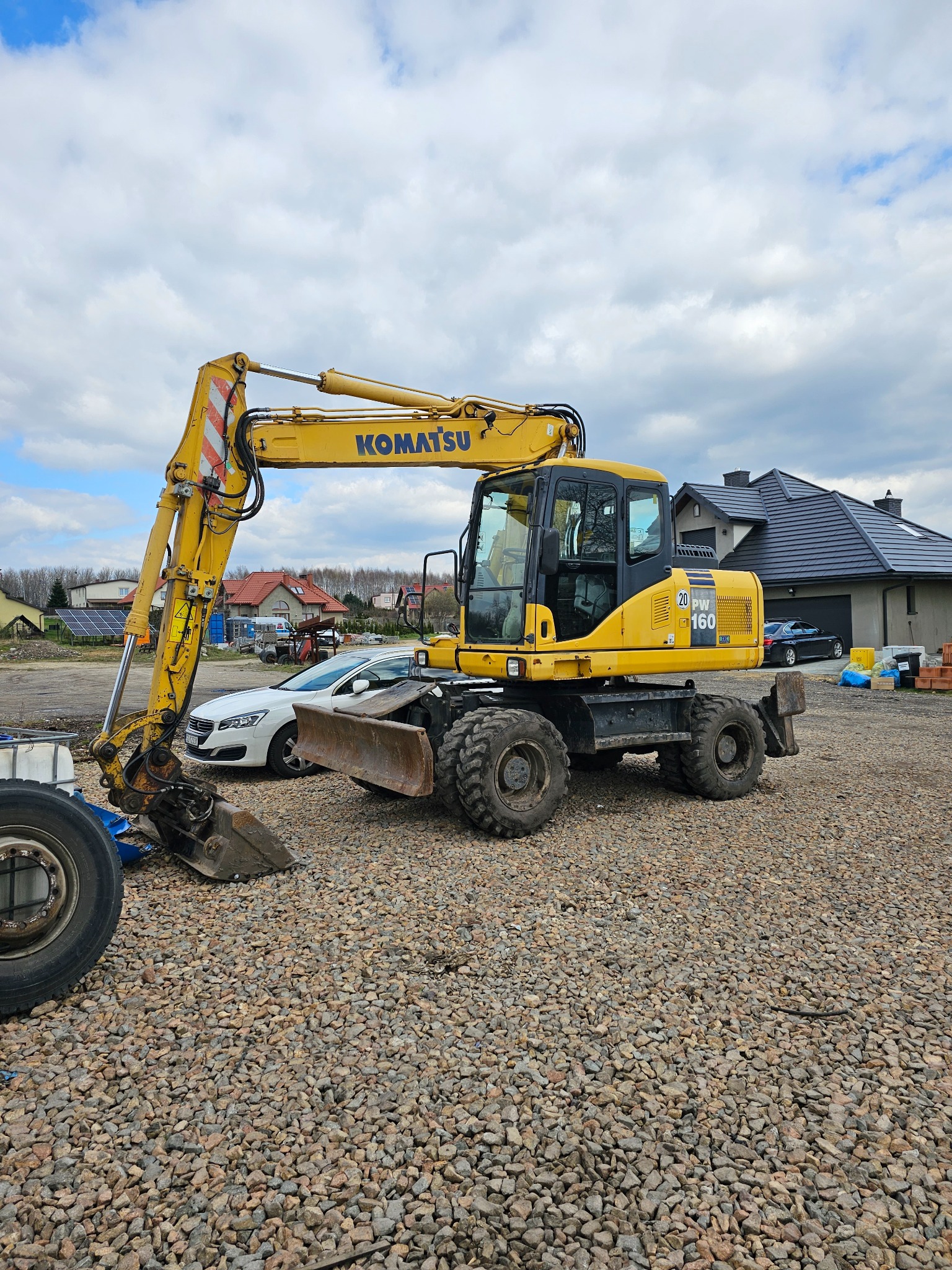 Żółta koparka kołowa Komatsu PW160 stoi na żwirowym placu budowy z widoczną łyżką i lemieszem, w tle biały samochód osobowy i dom z ciemną dachówką.