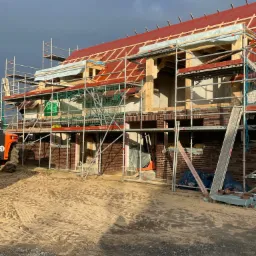 Budowa domu jednorodzinnego w trakcie: widoczne rusztowania, ściany z czerwonej cegły, drewniana konstrukcja dachu z czerwoną membraną dachową, burzowe chmury w tle.